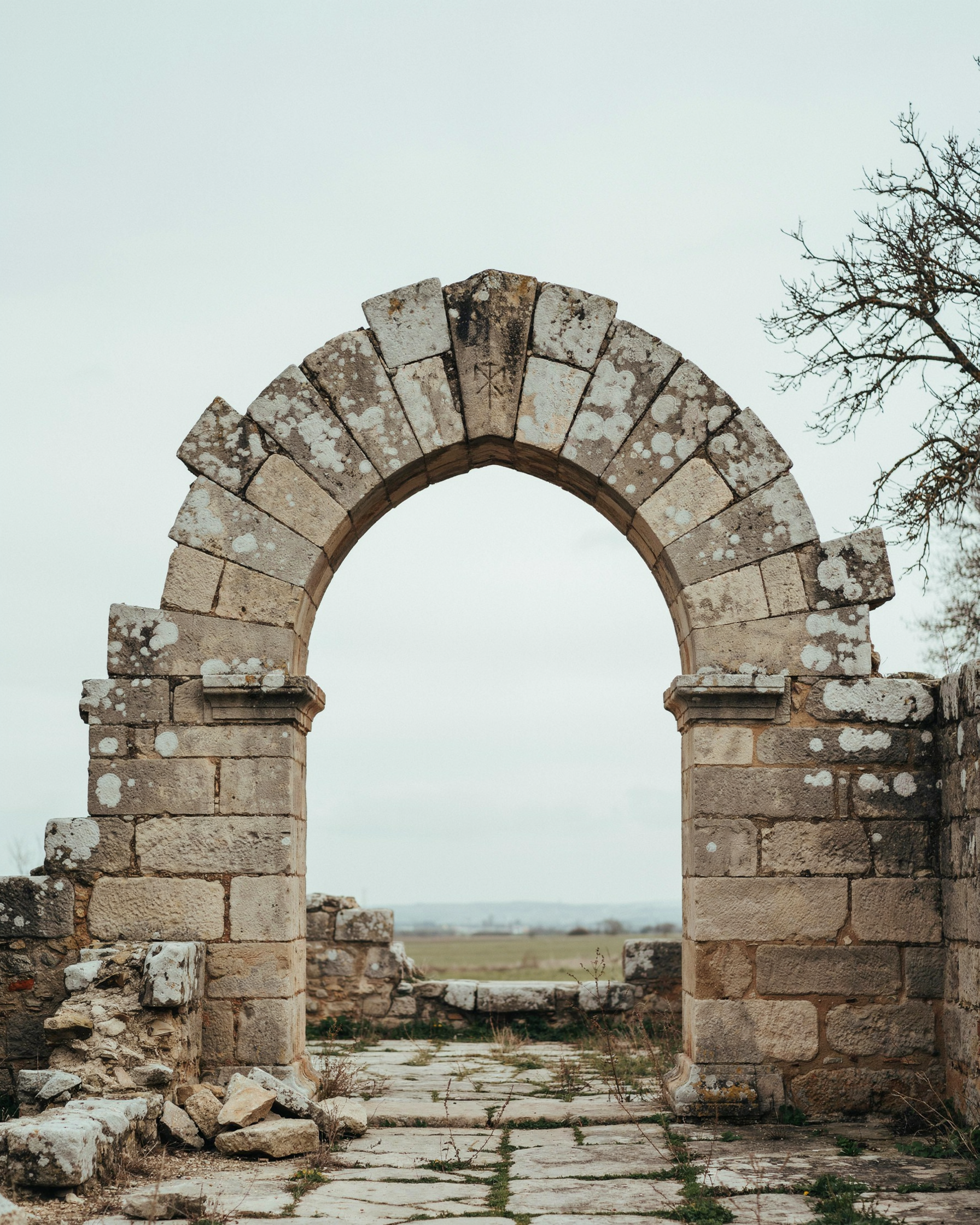 Editorial photograph of an old stone archway with one visible keystone at its apex, weathered masonry, flat overcast light, muted grey and sand tones, no people, no text, no branding, quiet and structural.