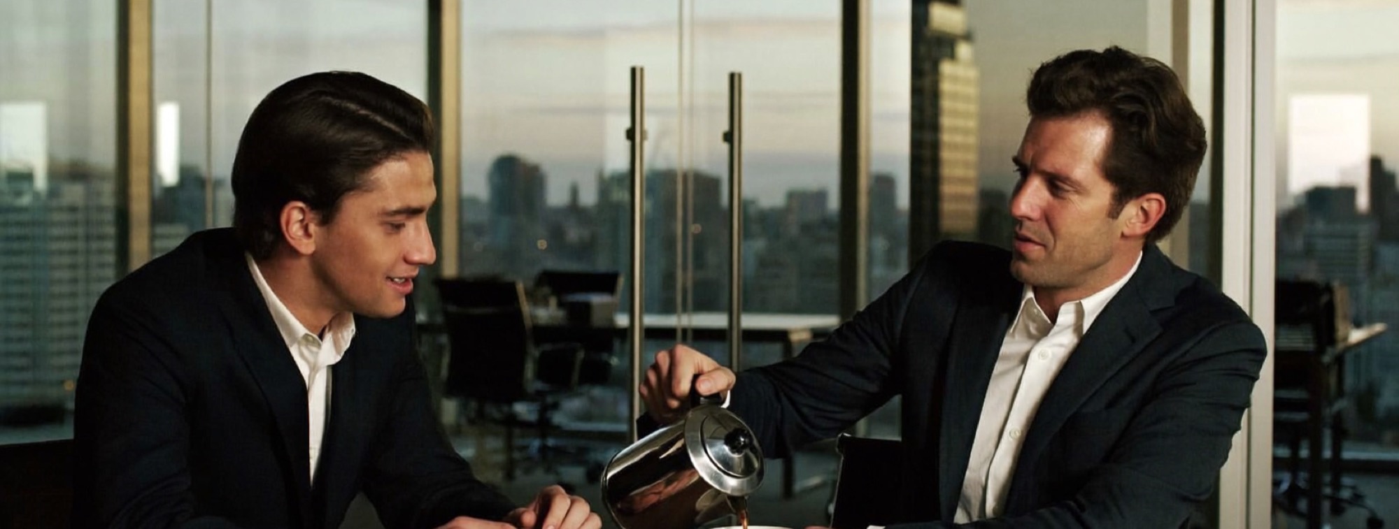 Two colleagues seated at a boardroom table at the end of the day, one pouring the other a coffee