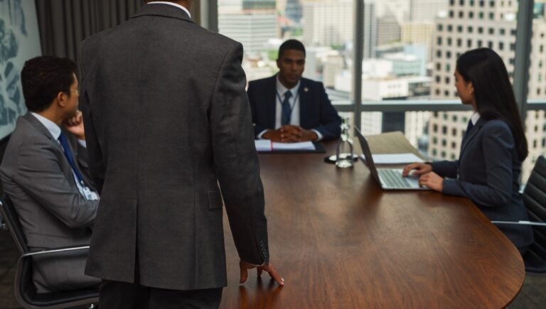 A consultant standing at the head of a boardroom table, three executives listening